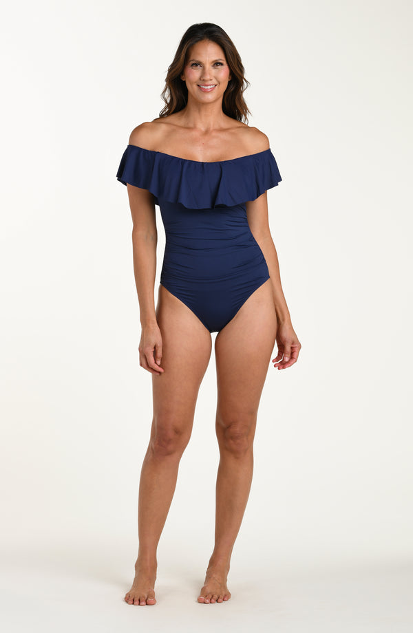 Full body front image of model wearing an indigo (navy blue) colored off-the-shoulder one-piece swimsuit featuring a flounce ruffle neckline and ruched bodice. 