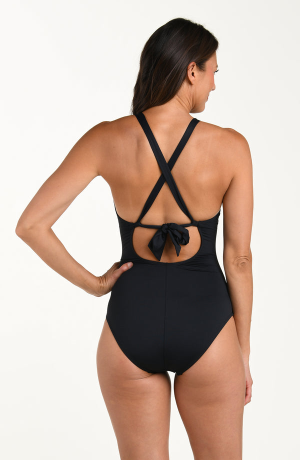 Back image of model wearing a solid black colored one-piece swimsuit with crisscross straps and a tie-back detail above a small cutout, and moderate rear coverage.