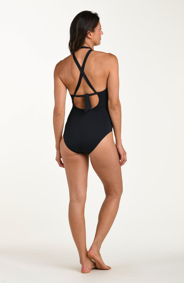 Full body back image of model wearing a solid black colored one-piece swimsuit with crisscross straps, tie-back detail above a small cutout, and moderate rear coverage.