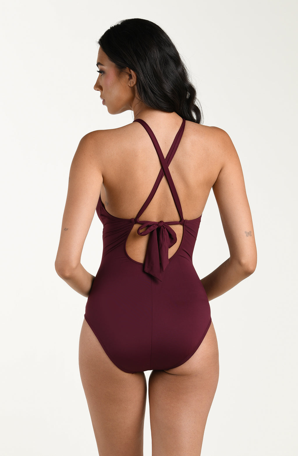 Back image of model wearing a dark cherry colored one-piece swimsuit with crisscross straps and a tie-back detail above a small cutout, and moderate rear coverage.