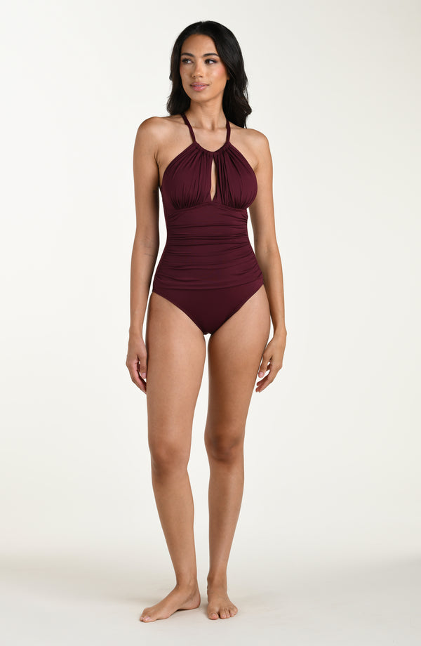 Full body front image of model wearing a dark cherry colored one-piece swimsuit with a halter neckline, keyhole cutout, and ruched detailing at the waist.