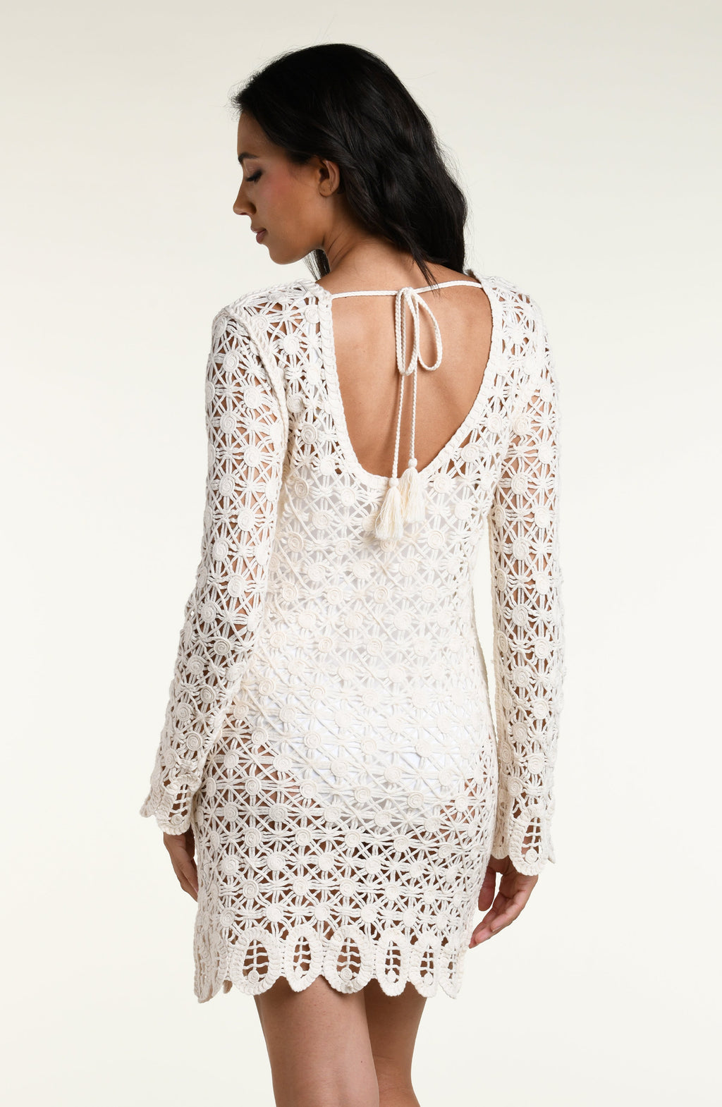 A woman modeling a white long-sleeve crochet mini dress with a scalloped hem against a plain light background.