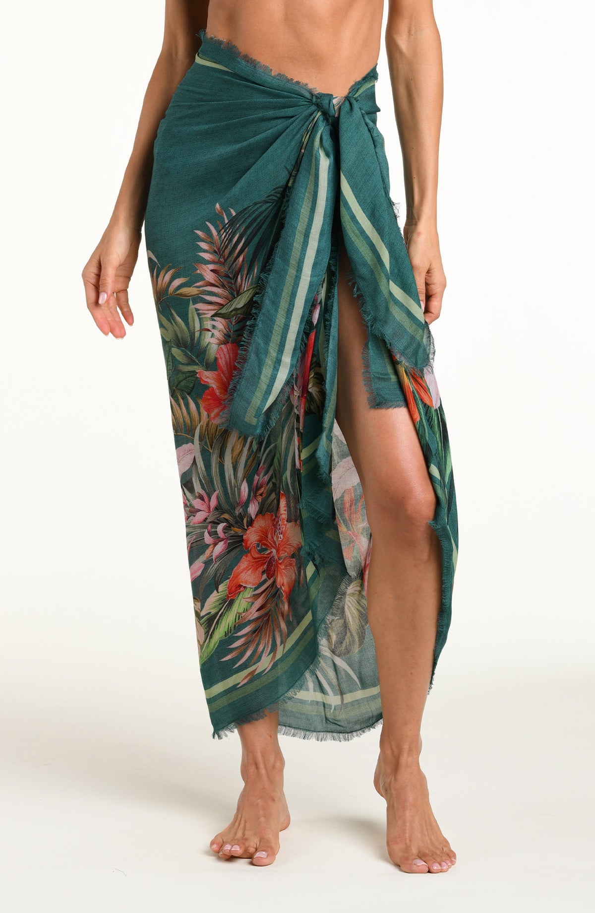 A model wearing a dark teal green tropical printed canga wrap skirt with a side slit, shown from the waist down against a white background.