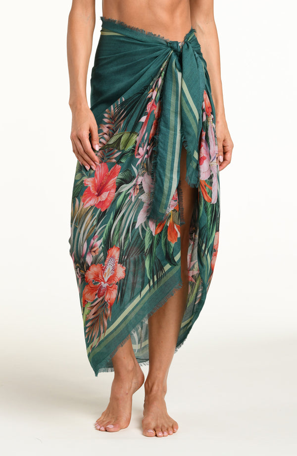 A model wearing a dark teal green tropical printed canga wrap skirt with a side slit, shown from the waist down against a white background.