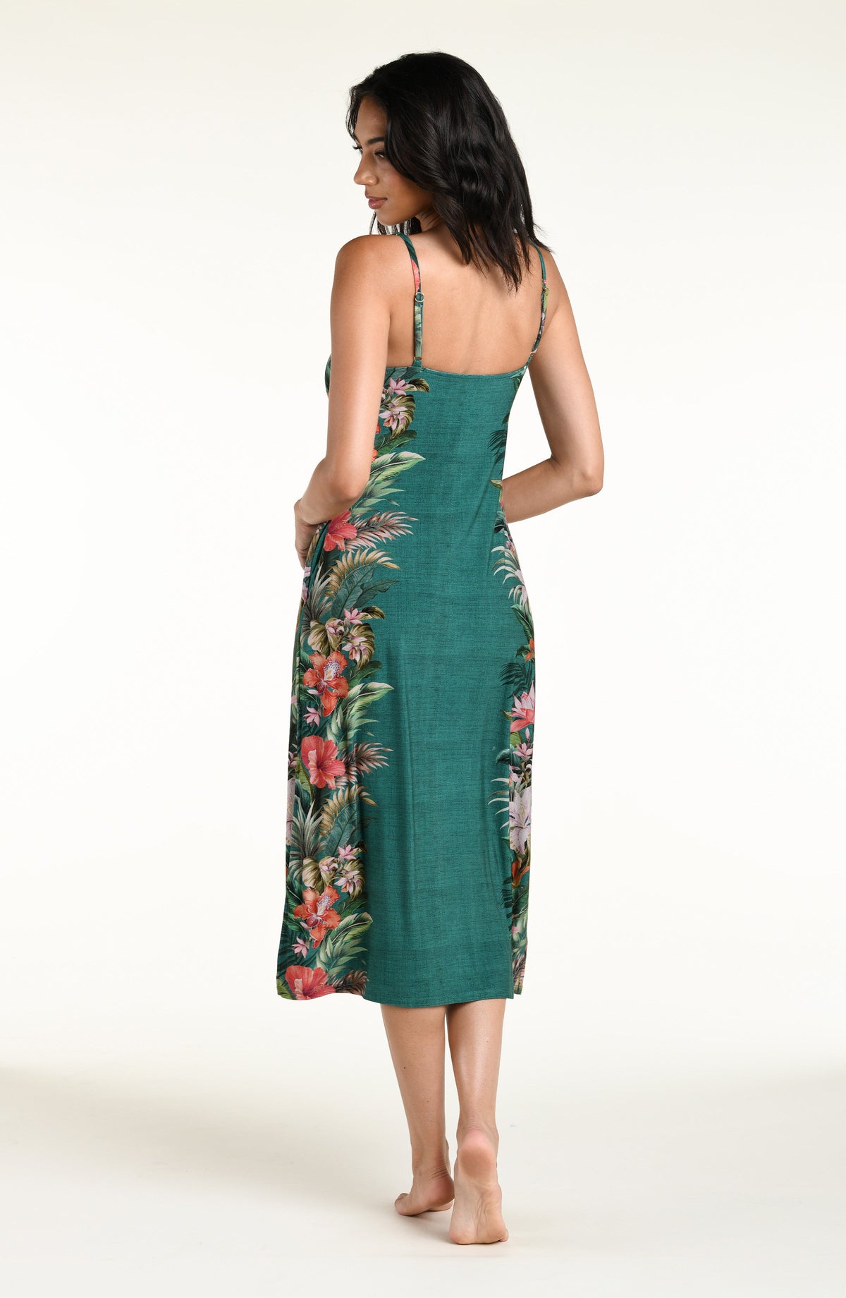A woman modeling a sleeveless midi dress from the back, featuring a teal green base with a vibrant tropical floral print in red, pink, and green, set against a plain white studio background.