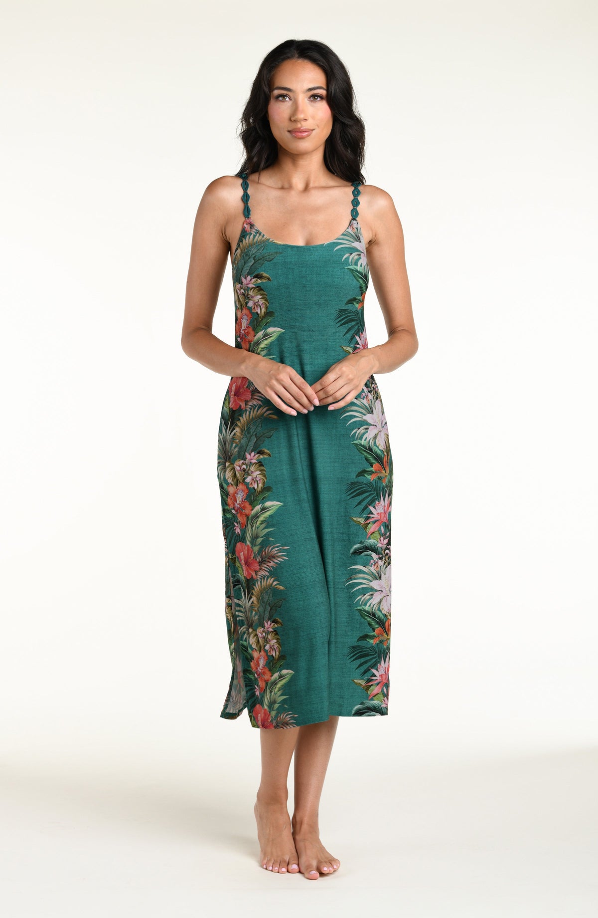 A woman modeling a sleeveless midi dress with thin straps and a green tropical floral print, standing against a plain white studio background.