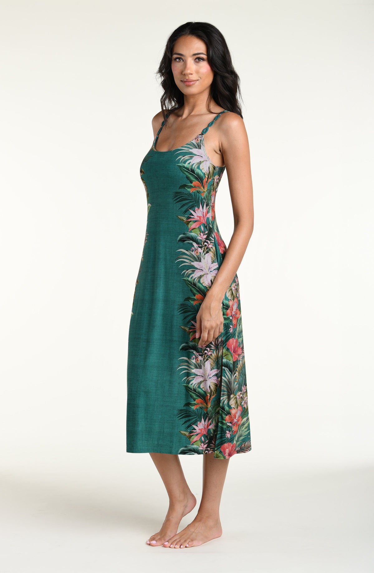 A woman modeling a sleeveless midi dress with thin straps and a tropical floral print in green, pink, orange, and white, photographed against a plain light background.