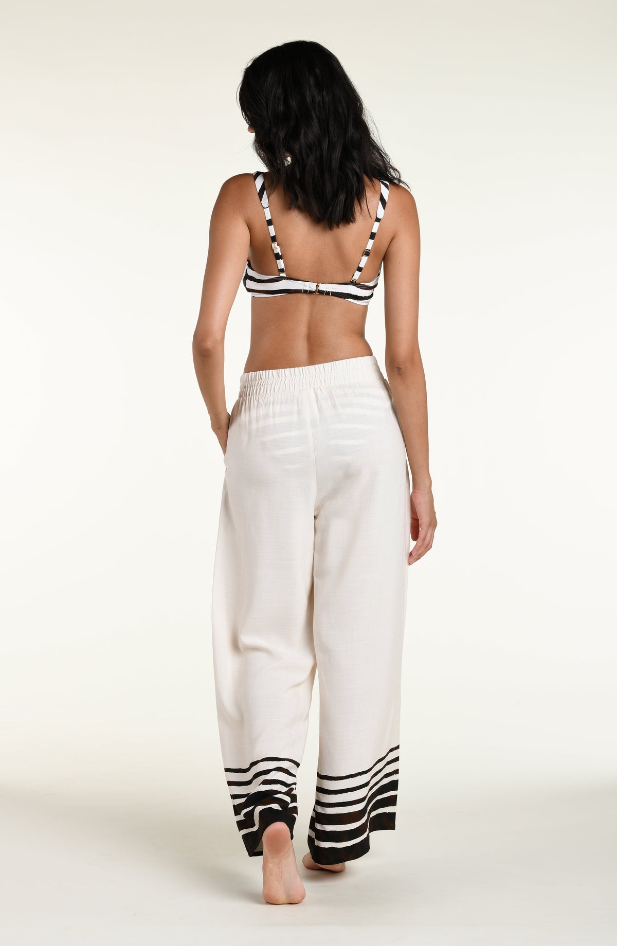 A woman modeling a brown and white striped bikini top with a front tie, paired with matching wide-leg white pants with brown stripes at the hems, set against a plain light background.
