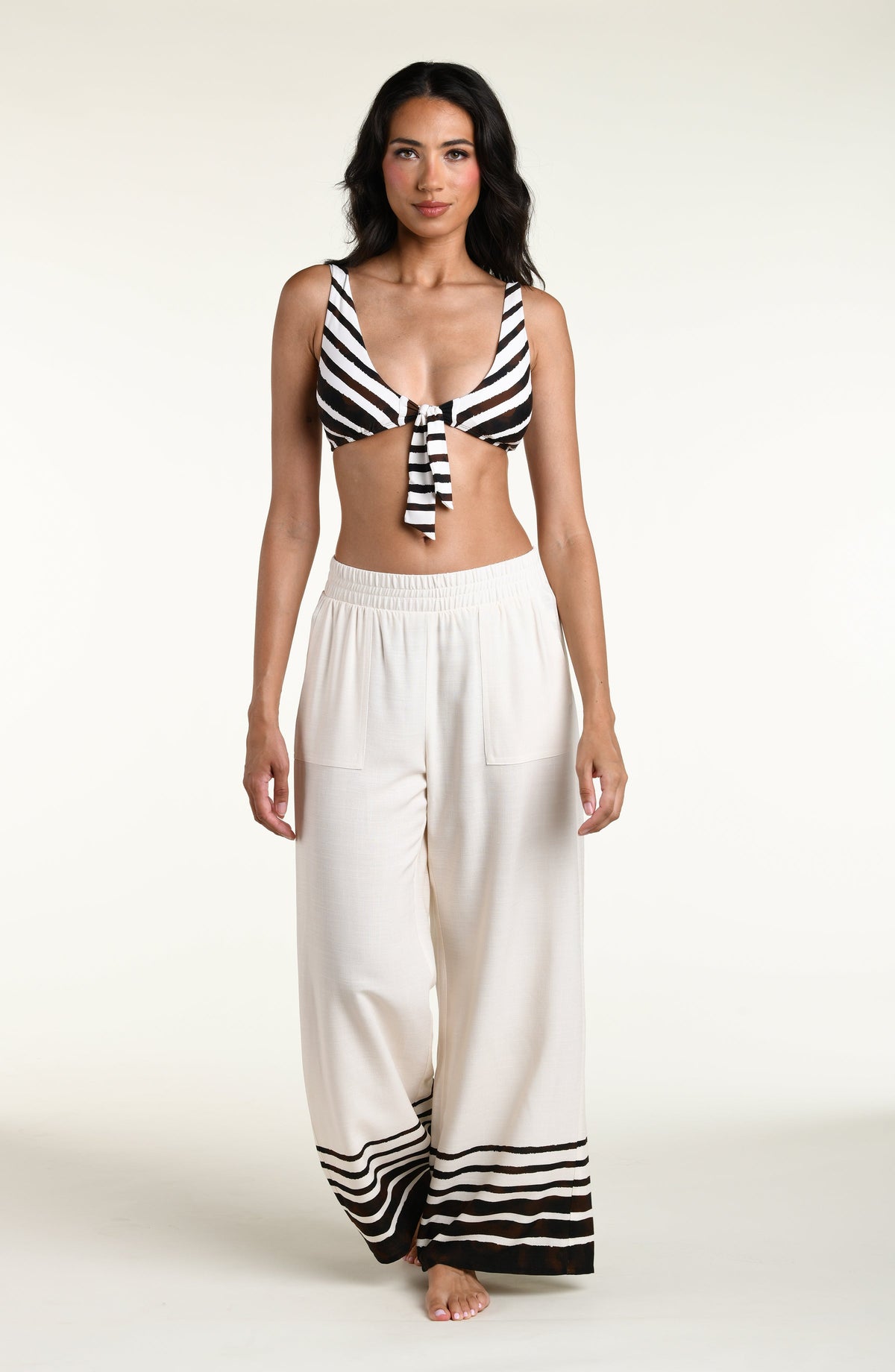 A woman modeling a brown and white striped bikini top with a front tie, paired with matching wide-leg white pants with brown stripes at the hems, set against a plain light background.