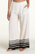 A woman wearing loose white wide-leg pants with brown horizontal stripes near the hem and a side pocket, shown against a plain light beige studio background.
