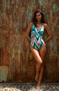 Woman wearing a teal multicolored one piece swimsuit against a rustic brown wall. 