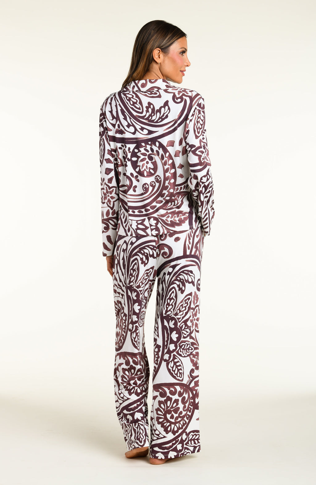 A woman modeling a matching long-sleeve button-front pajama top and wide-leg pajama pants set with an intricate paisley pattern in dark brown and white, set against a plain off-white studio background.