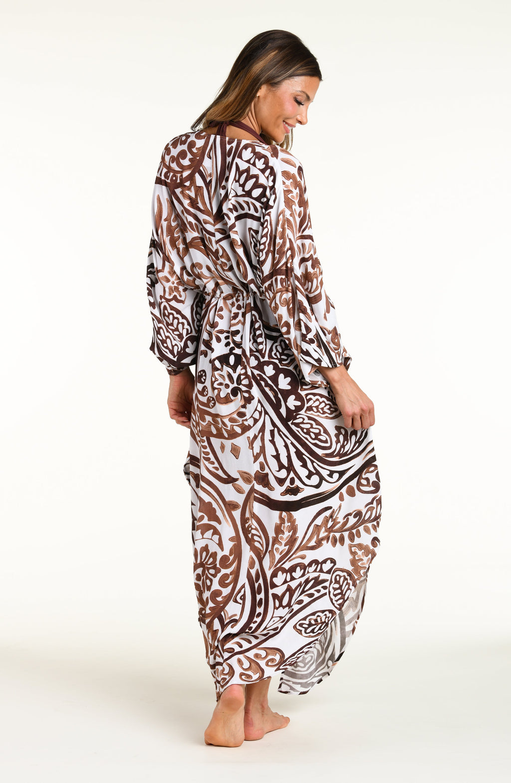 A woman modeling a long white caftan cover-up with a brown and white bohemian paisley pattern, standing against a plain white studio background.
