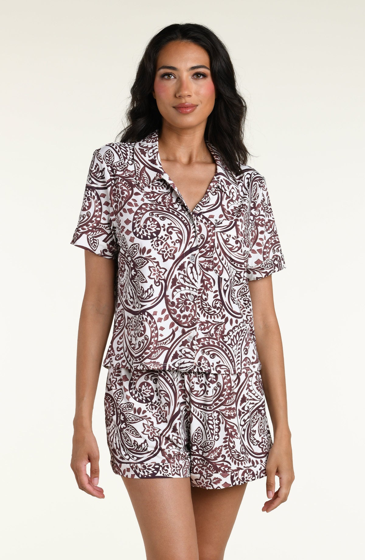 A woman modeling a pajama short-sleeve collared top and shorts set with an all-over brown paisley pattern on a white background, photographed against a plain light studio backdrop.