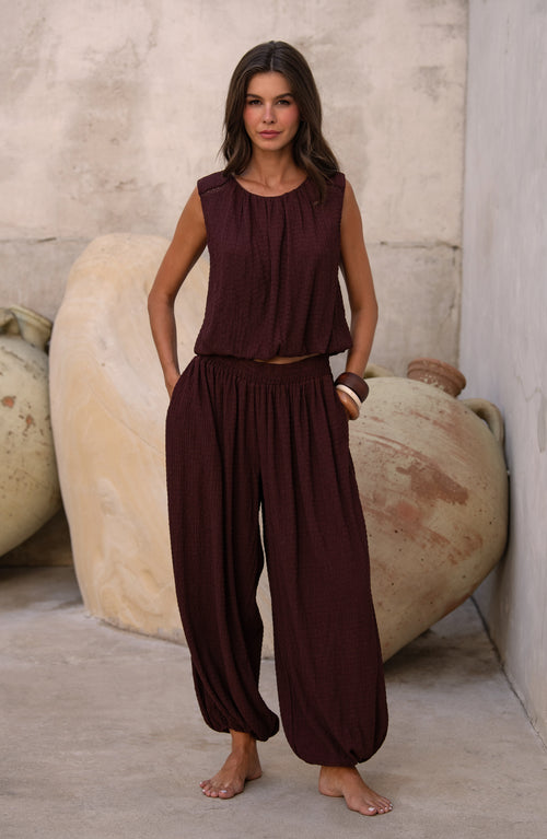 Woman wearing a java brown colored sleeveless top and matching pants against a light brown background. 