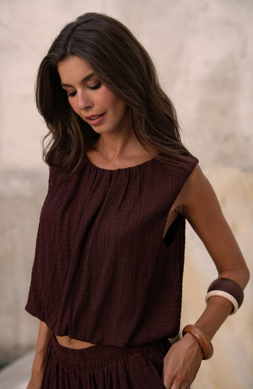 Woman wearing a java brown colored sleeveless top against a light brown background. 