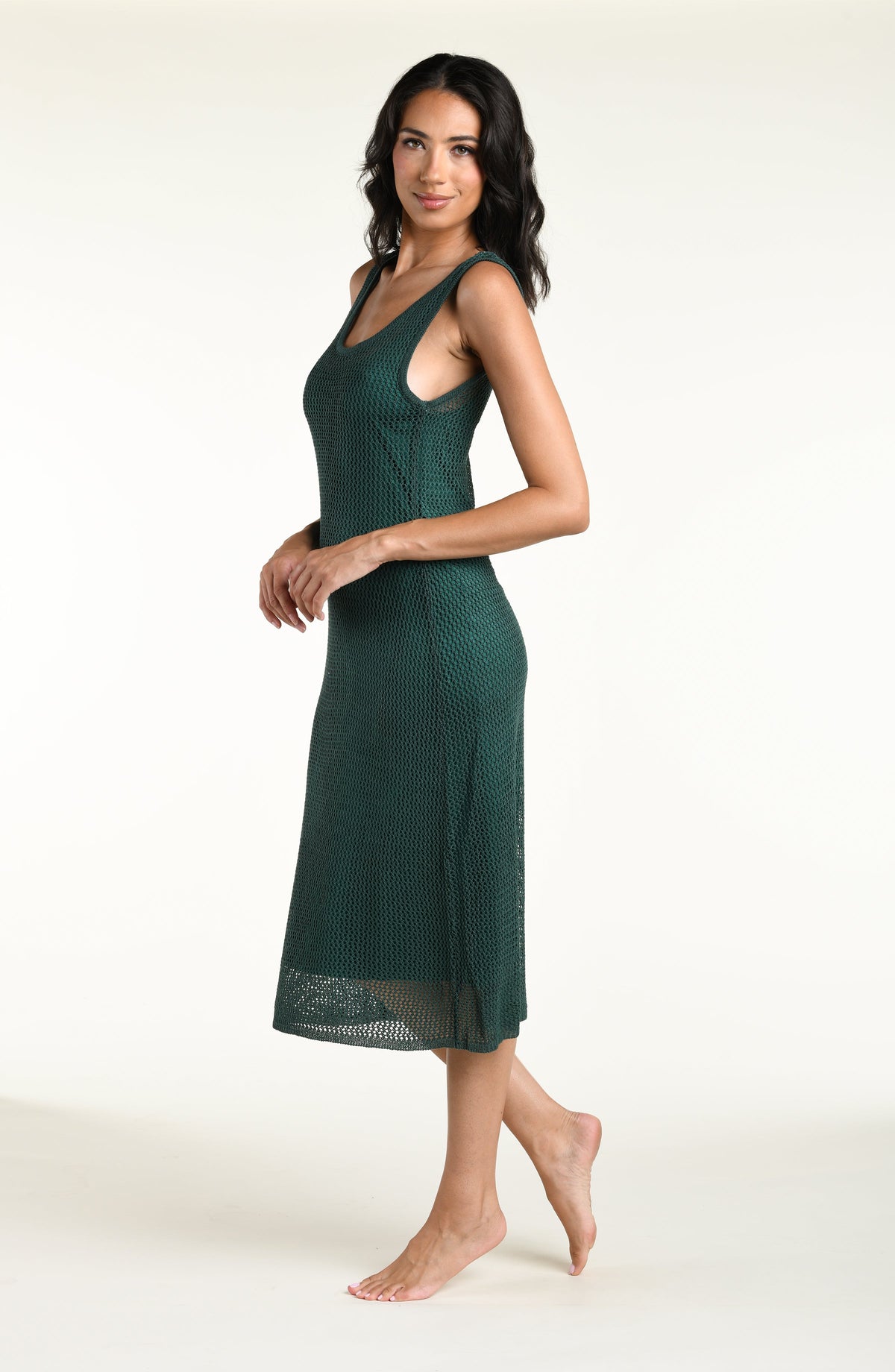 A woman modeling a dark green sheer mesh midi dress cover-up over a matching green bikini set, standing against a plain off-white studio background.