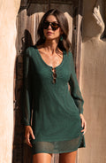 A woman modeling a dark green open-knit long-sleeve mini dress standing against a brown door. 