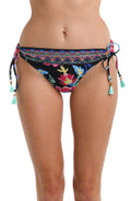 Front image of model wearing a black, pink, and blue multicolored floral patterned hipster bikini swimsuit bottom featuring a front band and side ties adorned with tassels.
