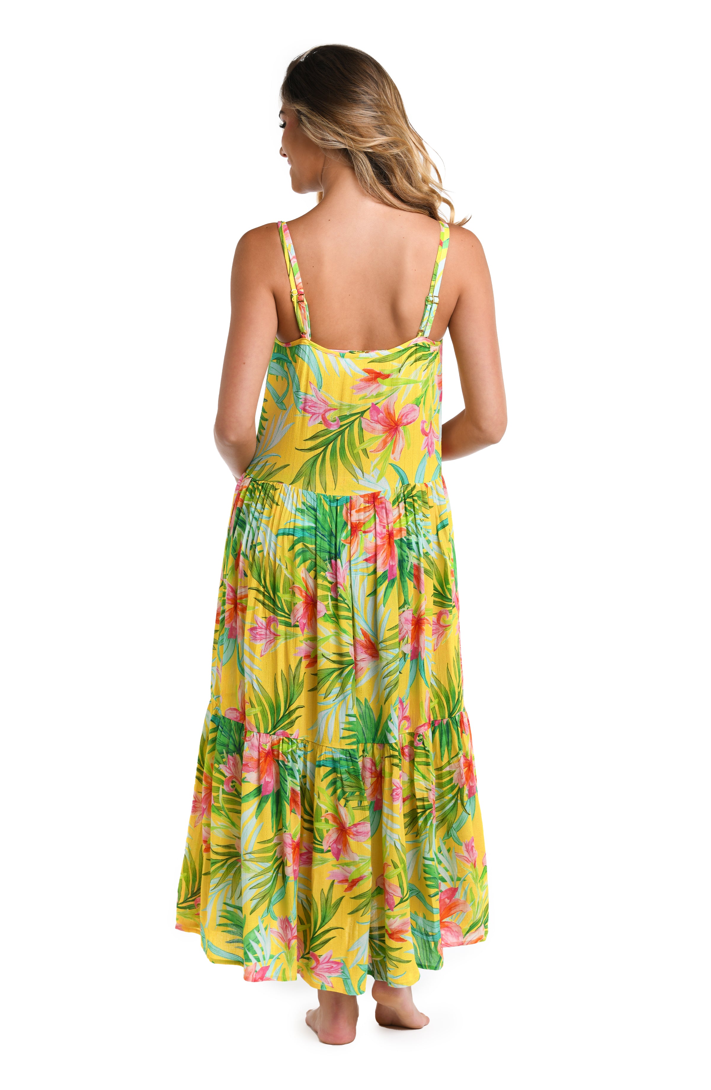 Back image of model wearing a bright yellow, pink, and green multicolored tropical printed midi dress featuring a scoop back, over the shoulder straps, pockets, and a tiered design.
