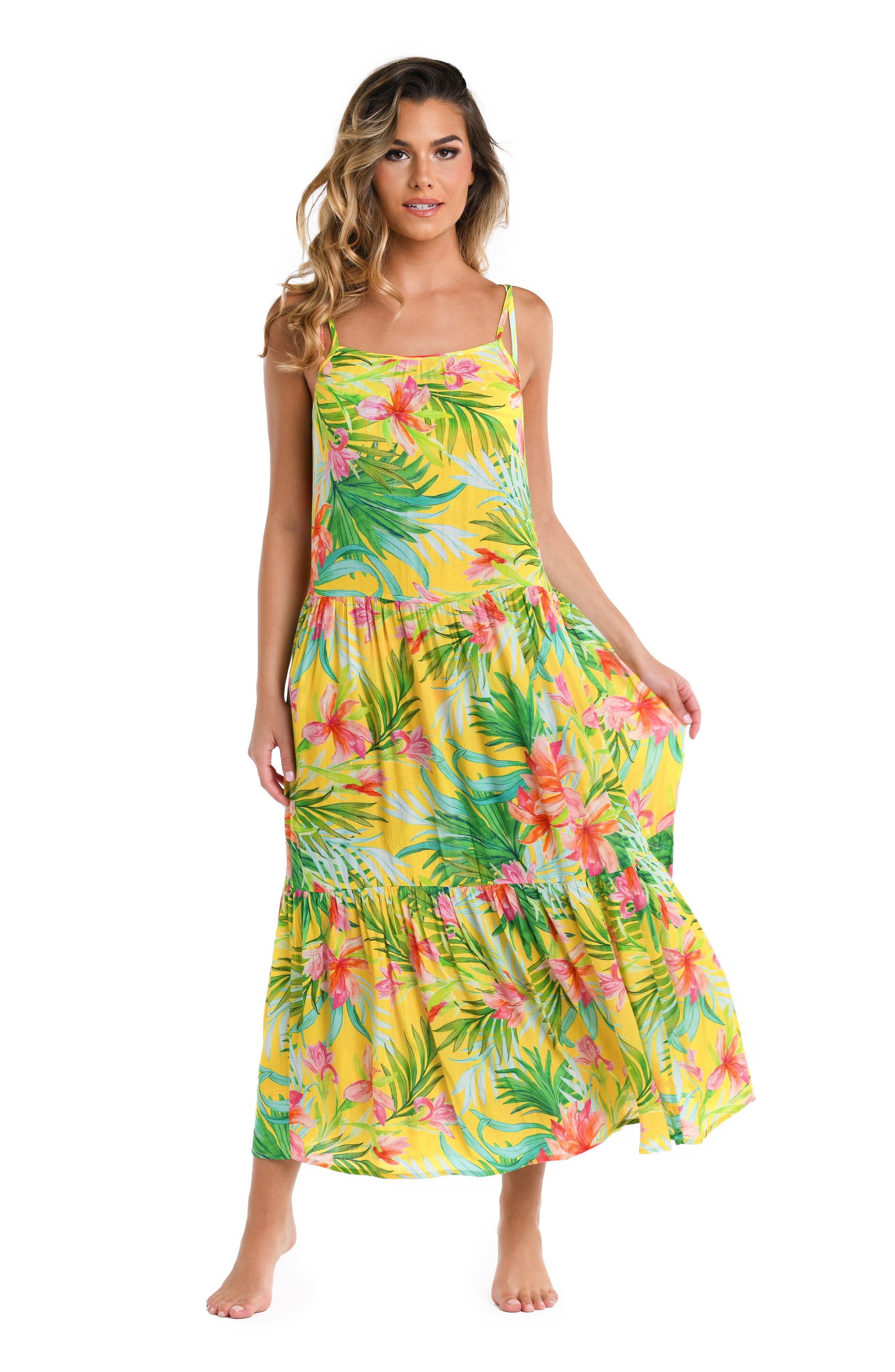 Front image of model wearing a bright yellow, pink, and green multicolored tropical printed midi dress featuring a scoop neckline, over the shoulder straps, pockets, and a tiered design.