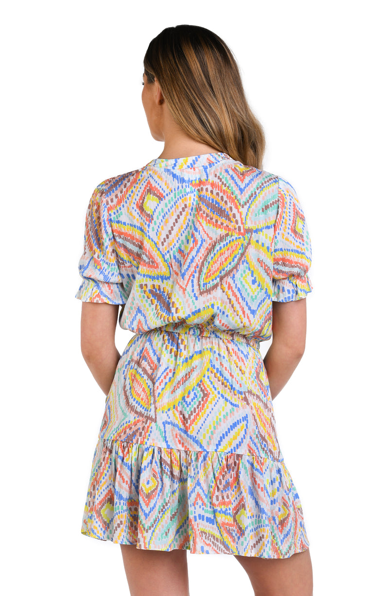 Model is wearing a geometric patterned cover-up dress in pastel shades of pink, blue, and yellow, accented with sporadic glimmers of gold sequin-like details on a white background.