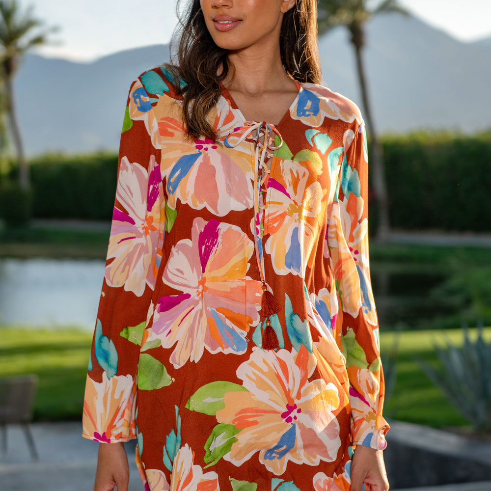 
                  
                    Model is wearing floral patterned tunic cover up with bold strokes of peach, pink, orange, and blue hues on a rich brown background.
                  
                