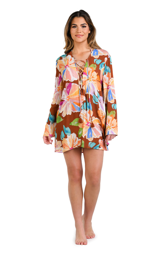 Model is wearing floral patterned tunic cover up with bold strokes of peach, pink, orange, and blue hues on a rich brown background.