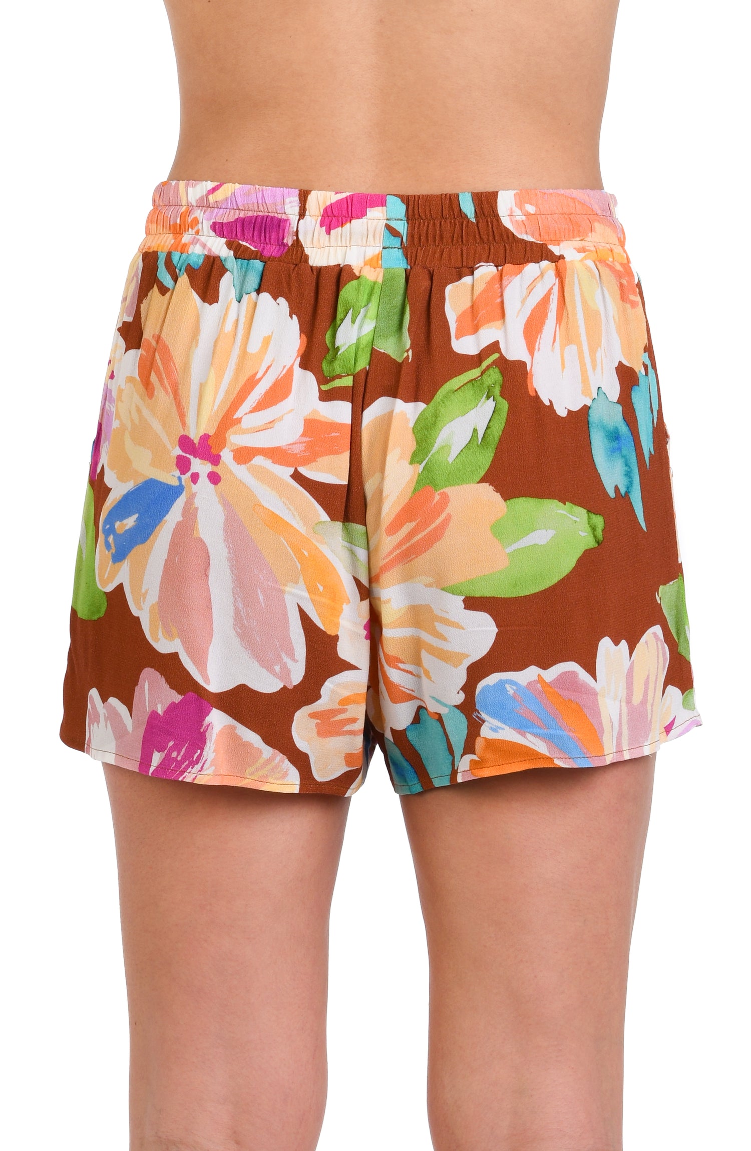 Model is wearing floral patterned beach short swimsuit cover up with bold strokes of peach, pink, orange, and blue hues on a rich brown background.