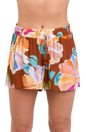 Model is wearing floral patterned beach short swimsuit cover up with bold strokes of peach, pink, orange, and blue hues on a rich brown background.