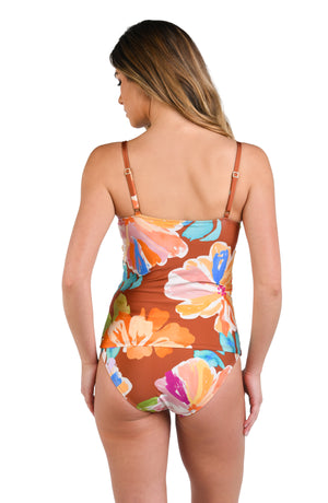 Model is wearing floral patterned swimsuit top with bold strokes of peach, pink, orange, and blue hues on a rich brown background.