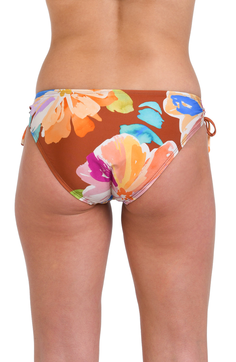 Model is wearing floral patterned swimsuit bottom with bold strokes of peach, pink, orange, and blue hues on a rich brown background.