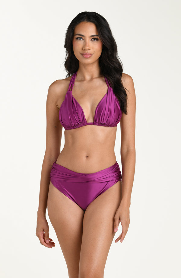 Full body front image of model wearing a halter bikini top in a berry color with ruched triangle cups and adjustable neck ties. This is paired with matching bikini bottoms. 