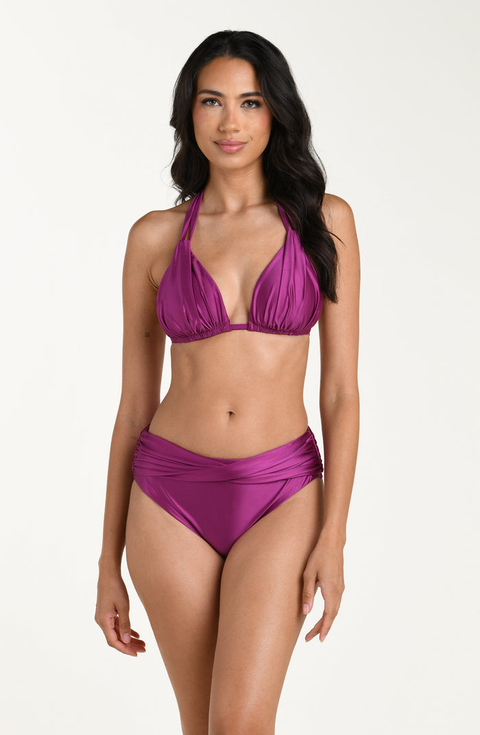 Full body front image of model wearing a halter bikini top in a berry color with ruched triangle cups and adjustable neck ties. This is paired with matching bikini bottoms. 