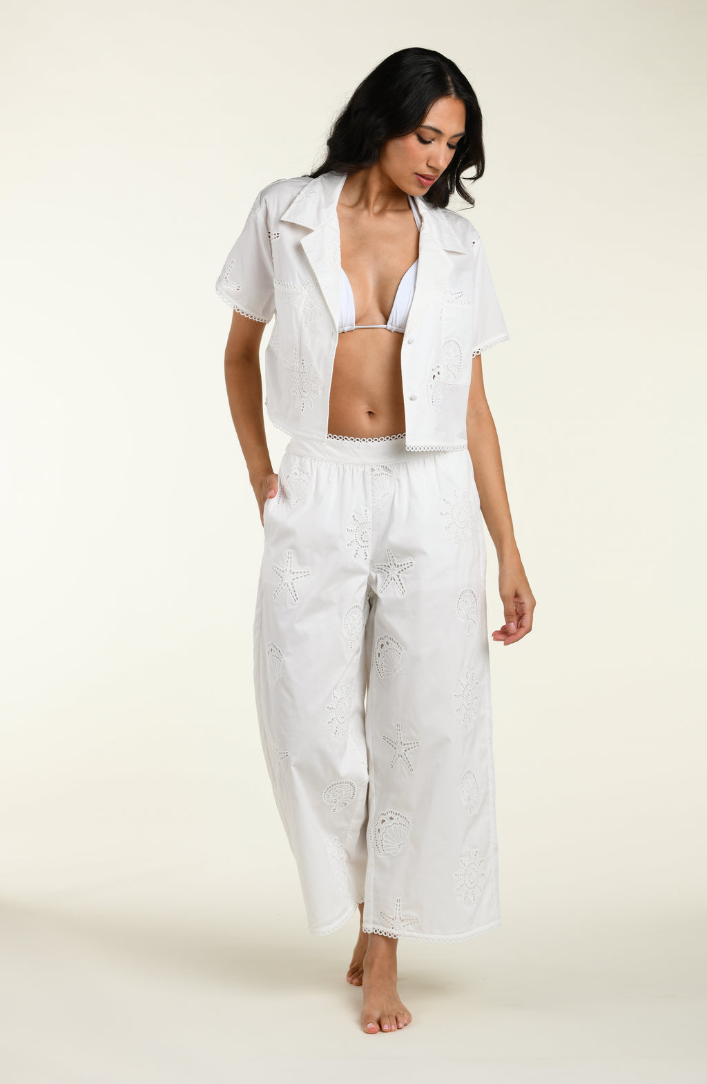Woman wearing a white cover up shirt with a white bikini top underneath, and matching pants against a white background. 