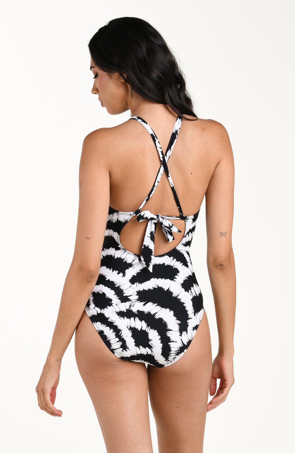 Back image of model wearing a black and white multicolored one piece swimsuit featuring a supportive, flattering fit with adjustable crisscross straps and a tie-back detail, and moderate rear coverage. 