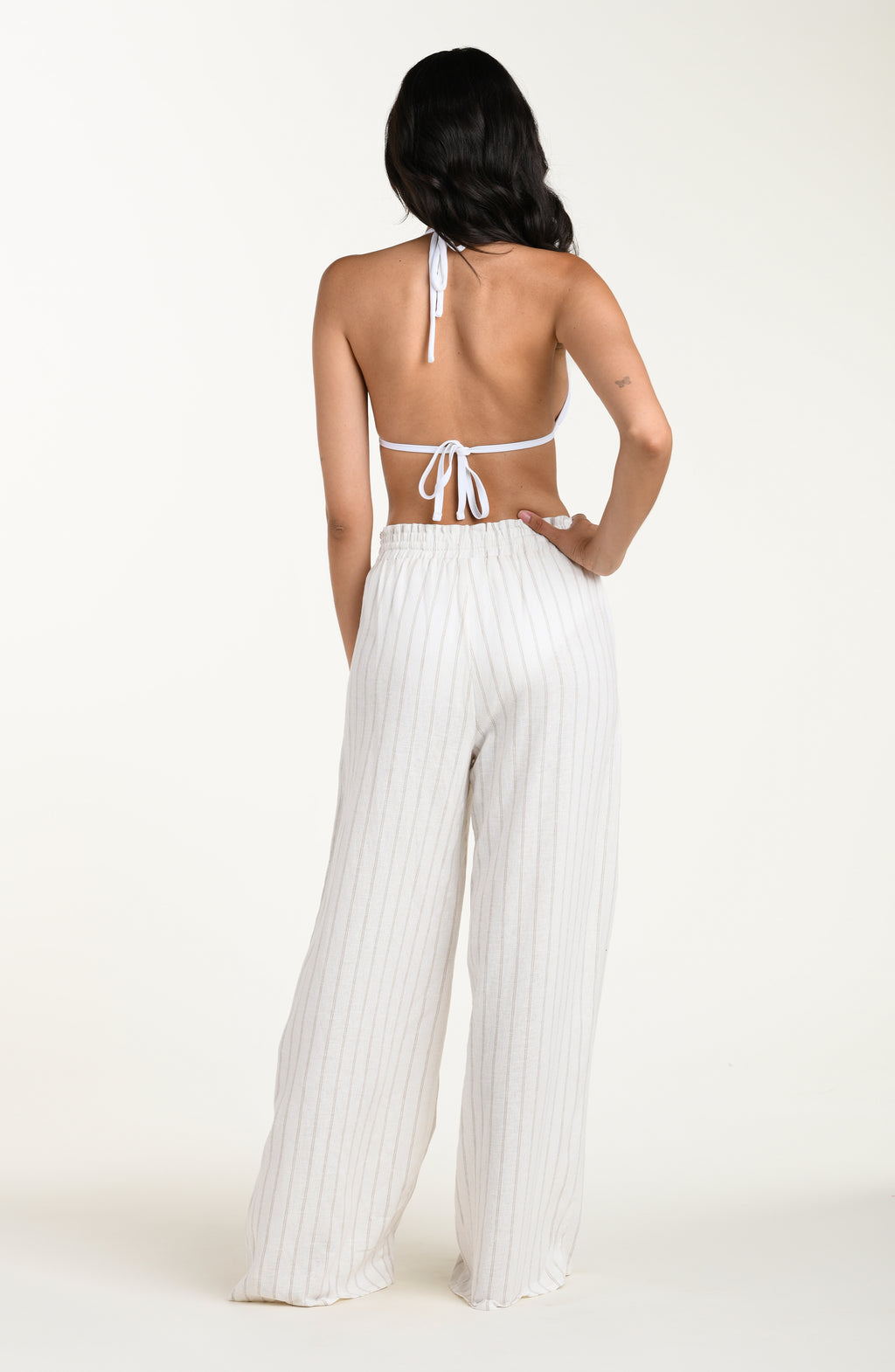 Full body back image of model wearing a cream striped colored wide-leg beach pants with a relaxed fit, drawstring waistband, and pockets. This is paired with a white bikini top. 