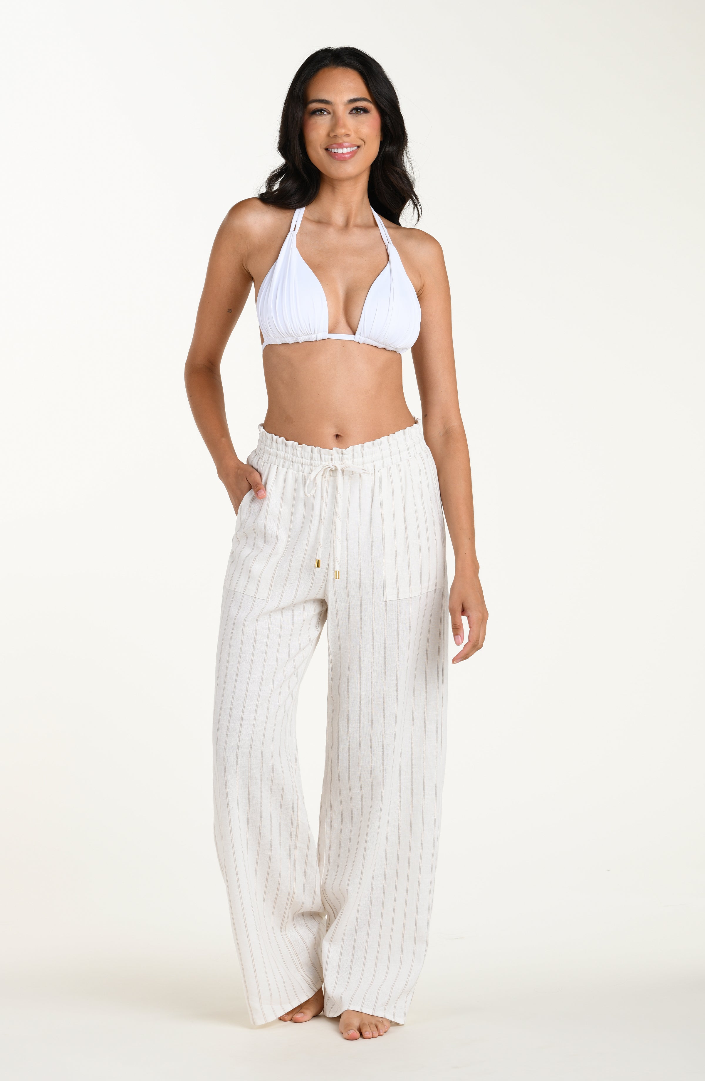 Full body front image of model wearing a cream striped colored wide-leg beach pants with a relaxed fit, drawstring waistband, and pockets. This is paired with a white bikini top.