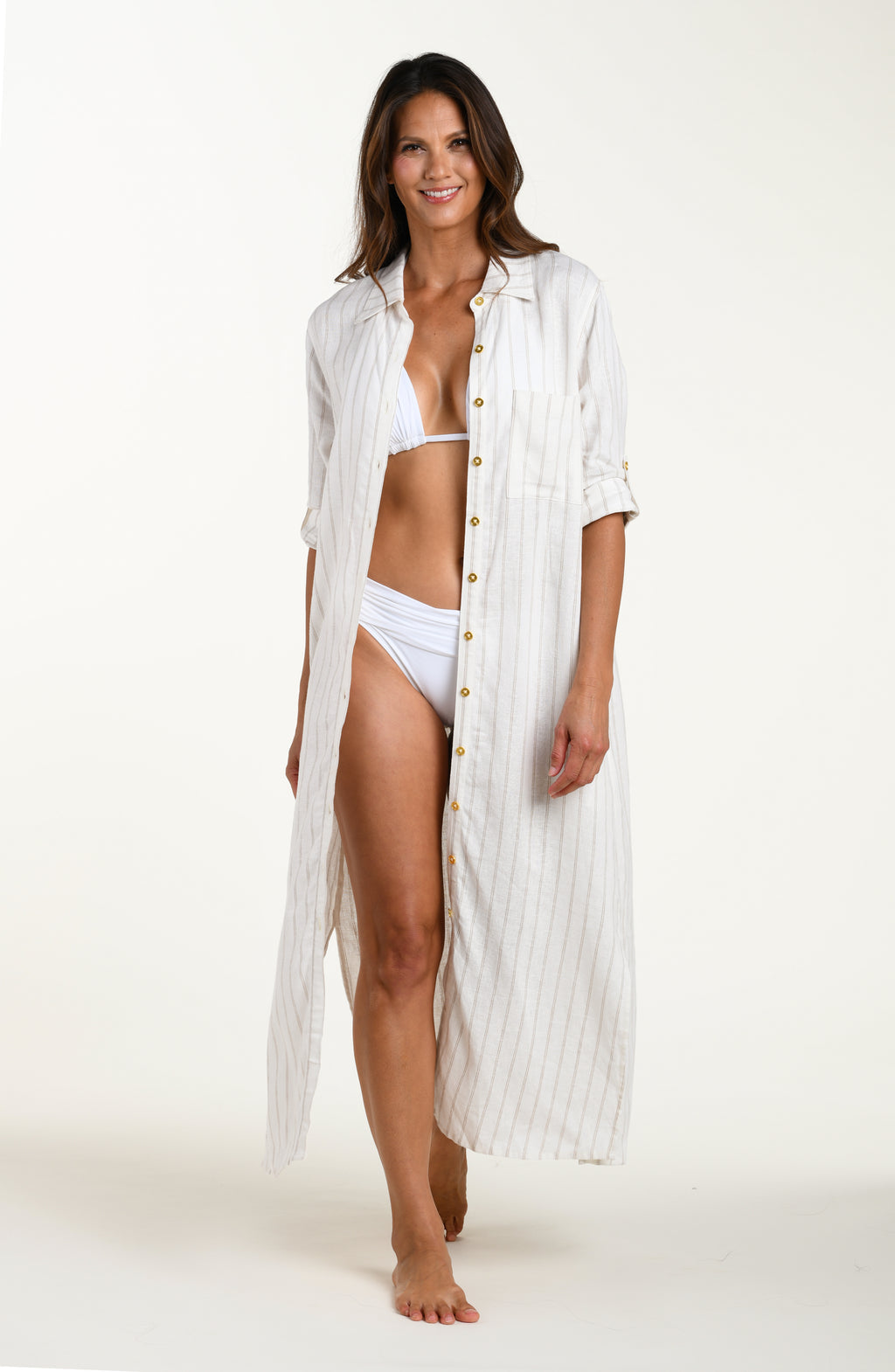Full body front image of model wearing a cream striped colored button-down, shirt dress cover-up with roll-tab sleeves, a belted waist, and side slits. Model is showcasing cover up open with a white bikini top and bottom underneath. 