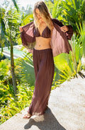 Woman in a brown outfit standing in a tropical setting with green foliage.