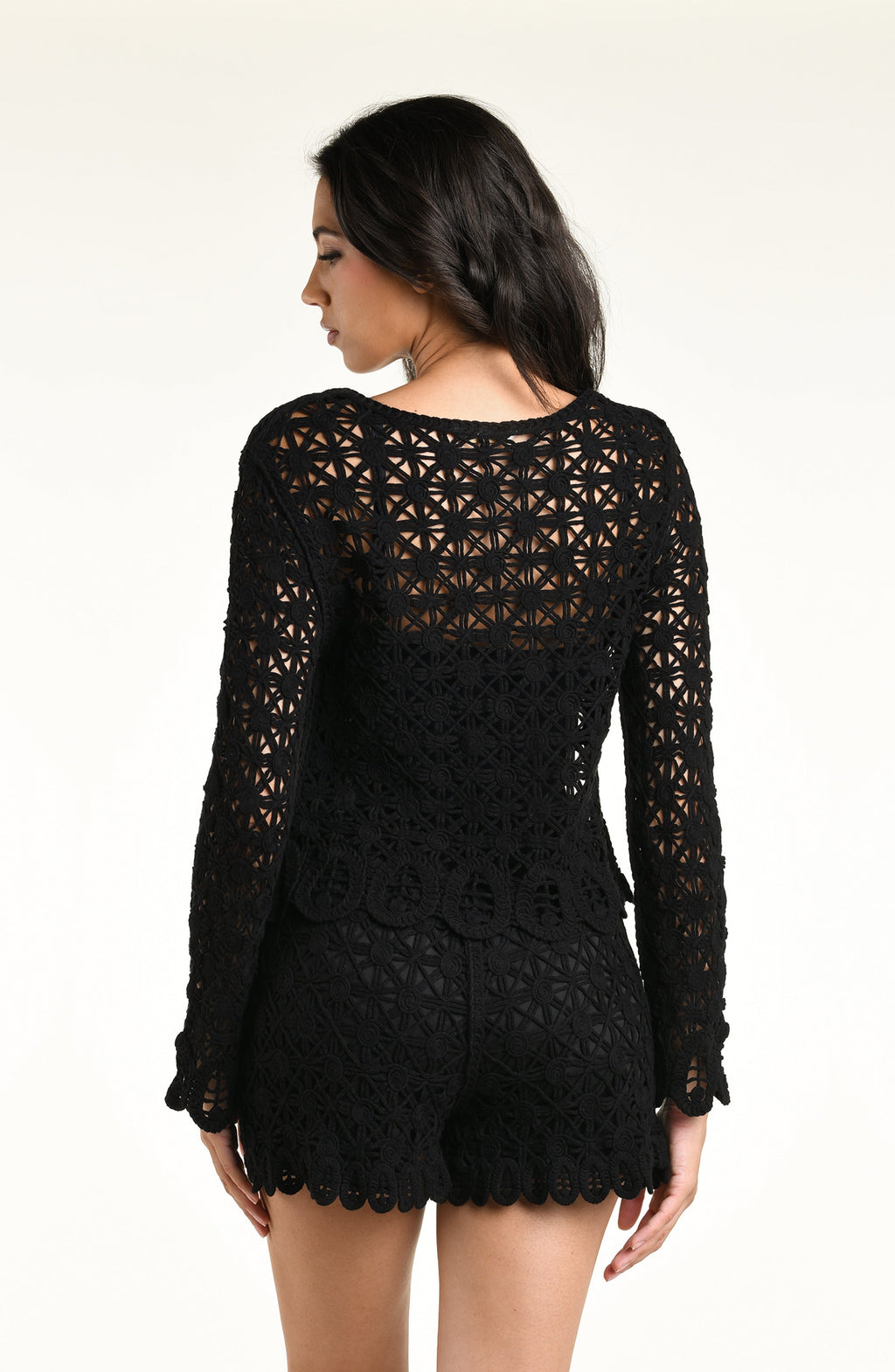 A woman modeling a black long-sleeve crochet top and matching short with a scalloped hem and tassel detail, set against a plain light background.