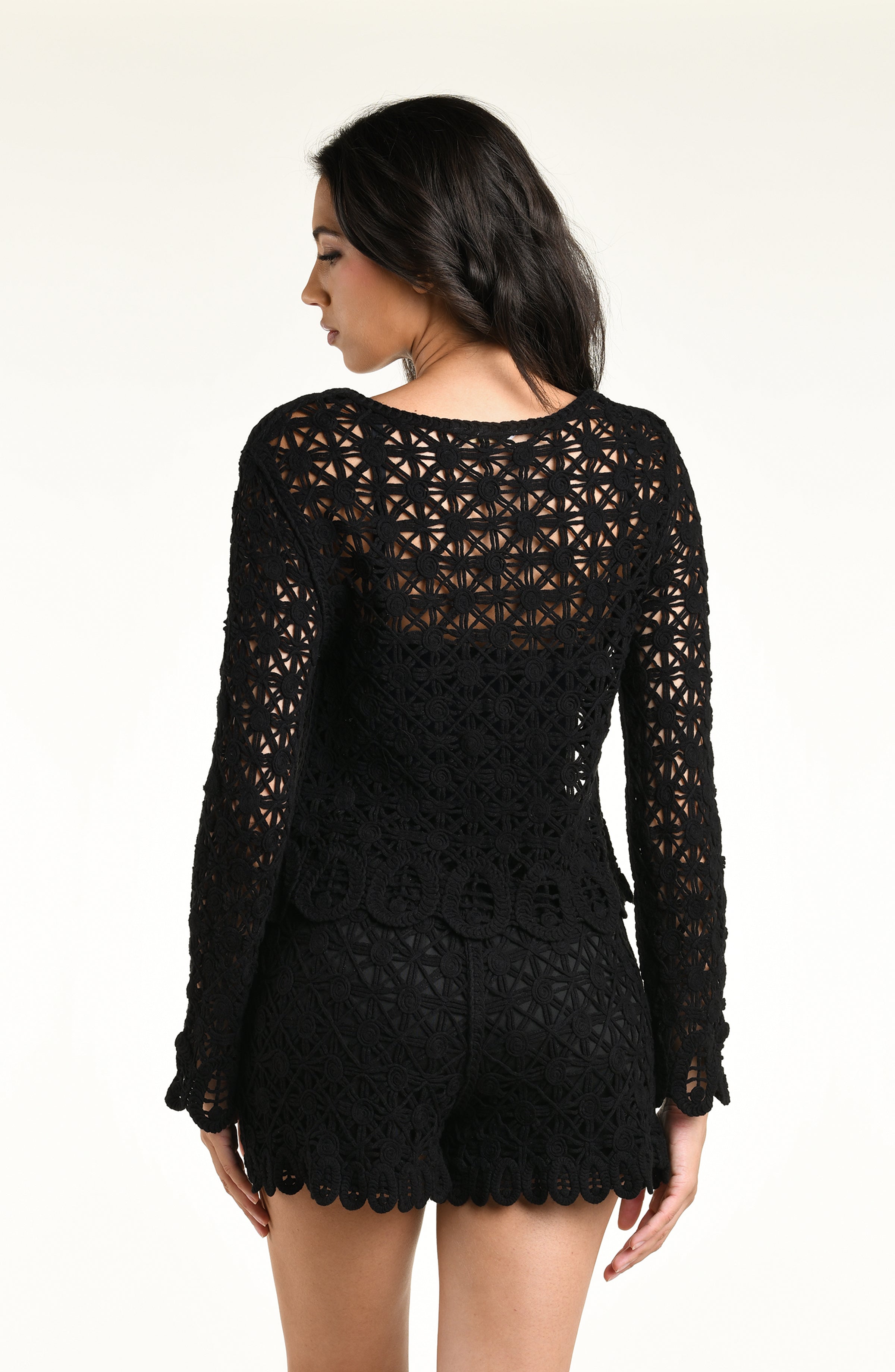 A woman modeling a black long-sleeve crochet top and matching short with a scalloped hem and tassel detail, set against a plain light background.