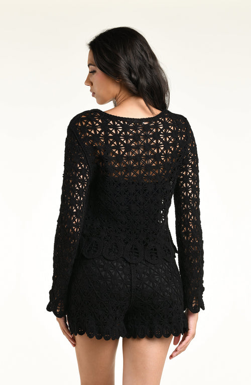 A woman modeling a black long-sleeve crochet top and matching short with a scalloped hem and tassel detail, set against a plain light background.