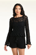 A woman modeling a black long-sleeve crochet top and matching short with a scalloped hem and tassel detail, set against a plain light background.