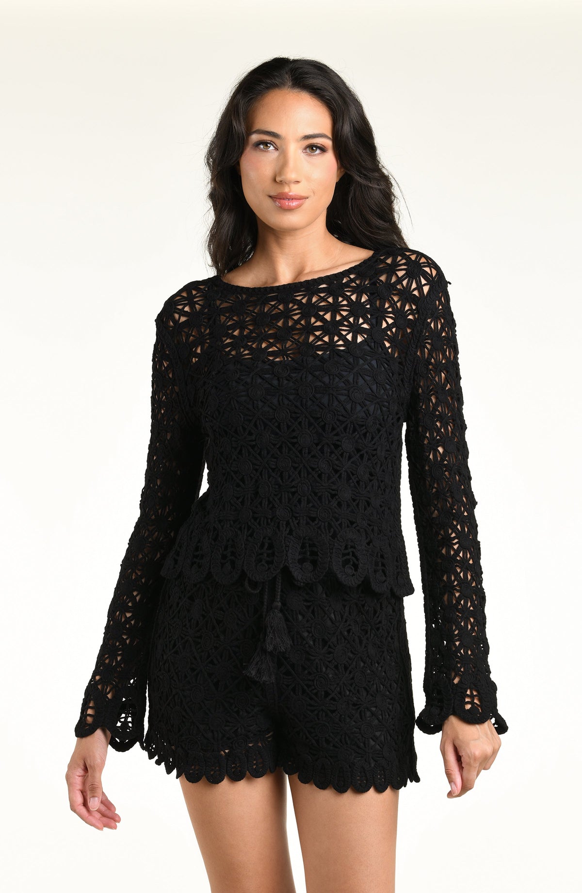 A woman modeling a black long-sleeve crochet top and matching short with a scalloped hem and tassel detail, set against a plain light background.