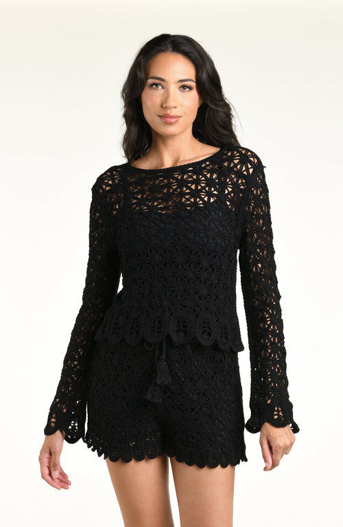 A woman modeling a black long-sleeve crochet top and matching short with a scalloped hem and tassel detail, set against a plain light background.