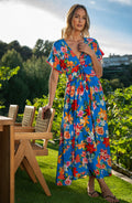 Woman in a colorful floral dress standing outdoors with greenery and a table in the background