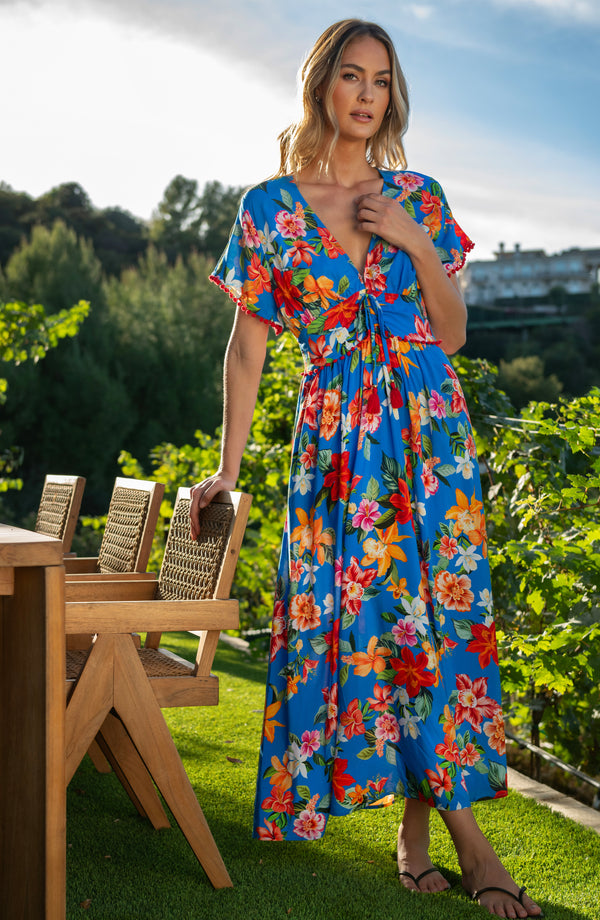 Woman in a colorful floral dress standing outdoors with greenery and a table in the background