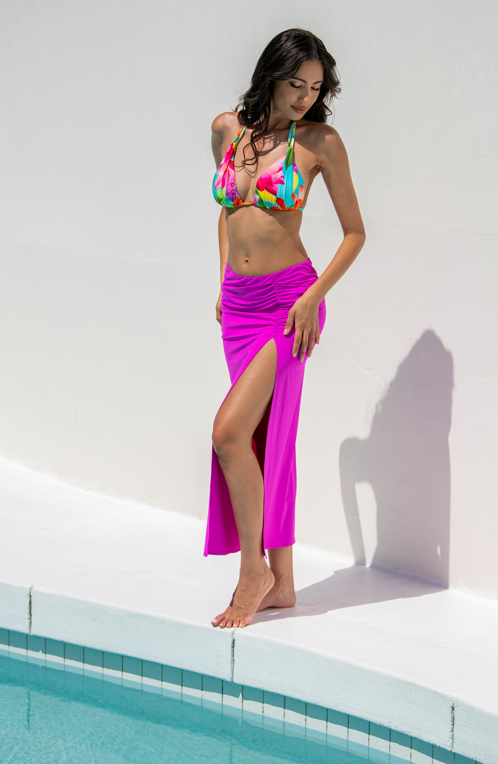 Woman in colorful floral bikini top and violet colored skirt standing by a pool.