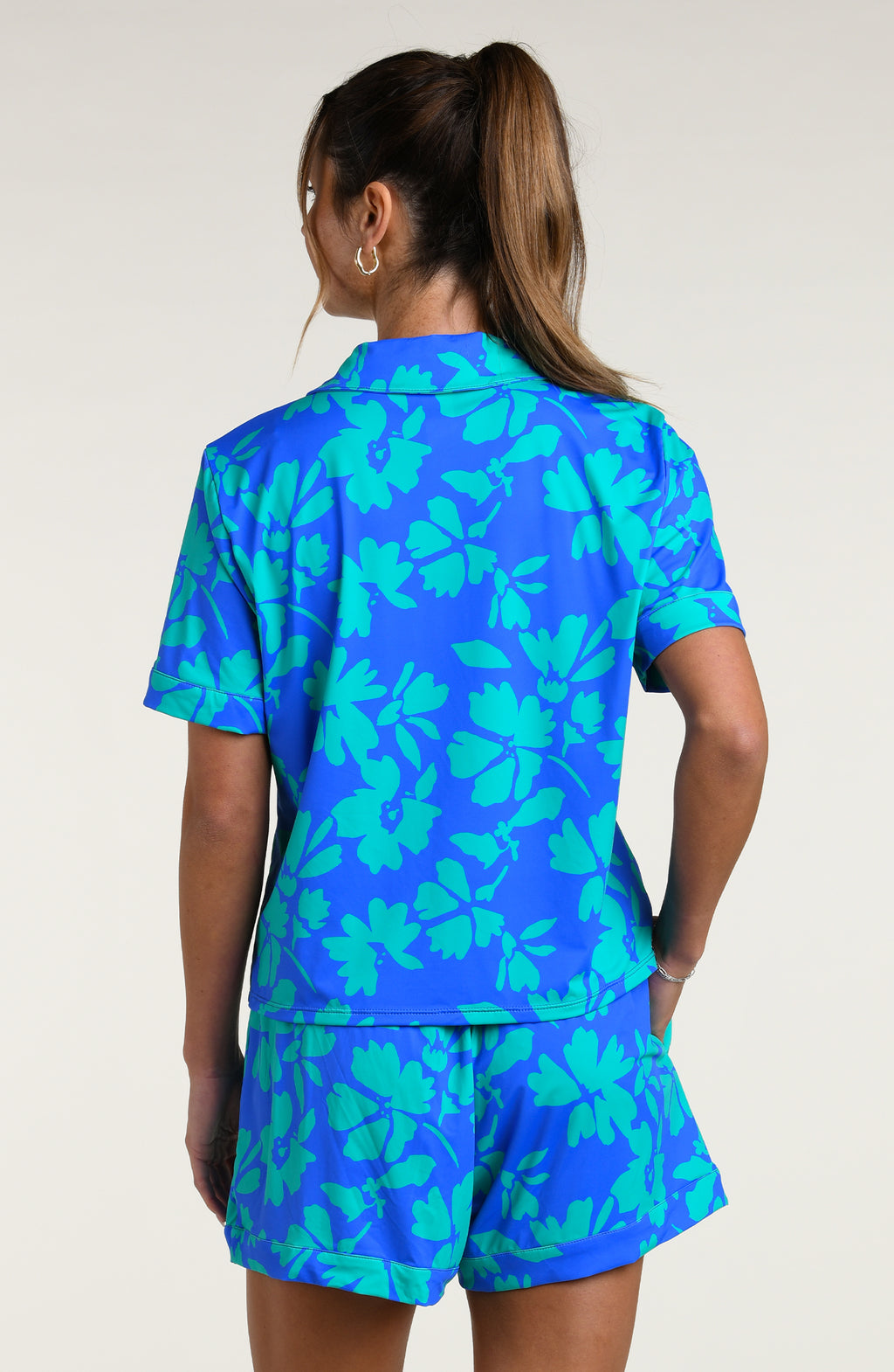 Woman wearing a blue and green floral patterned short sleeve shirt and shorts pajama set against a white background